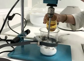 Stéphanie la créatrice, verse une huile dorée dans un bécher contenant une base blanche, lors de la préparation d’une émulsion cosmétique naturelle à l’aide d’un agitateur de laboratoire. L’image illustre le processus de formulation d’un déodorant bio et artisanal.